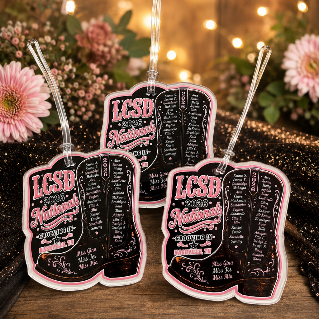 Three black and pink LCSD National tags with straws on a wooden surface with flowers and lights in the background.