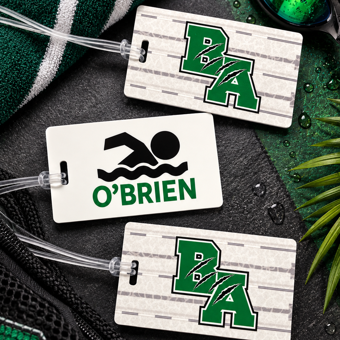Three custom luggage tags arranged on a dark textured surface with a green and white striped towel, swim goggles, and water droplets. Two tags feature a green and white wood-grain pattern with the letters 'BA' in green, and one tag is solid white with a black swim icon and the name 'O'BRIEN' in green.